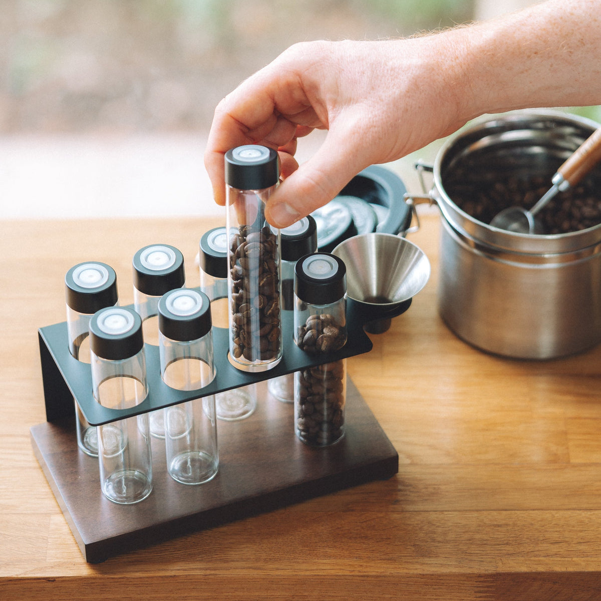 A hand holds a clear glass vial with a black lid containing coffee beans from a black plastic organizer tray on a wooden table, with a metal pot in the background.