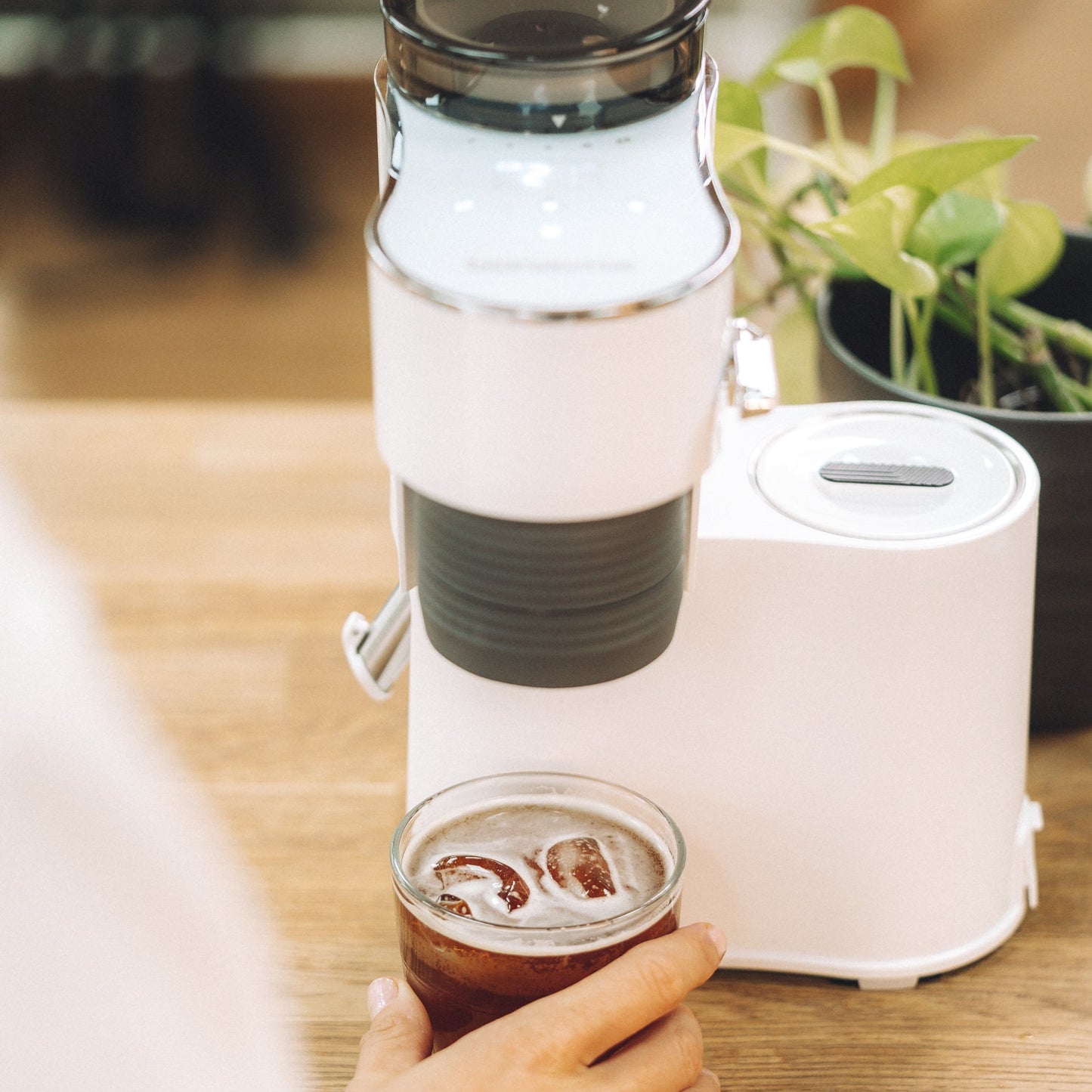 Person holding a glass of iced coffee next to a portable coffee maker on a wooden table.