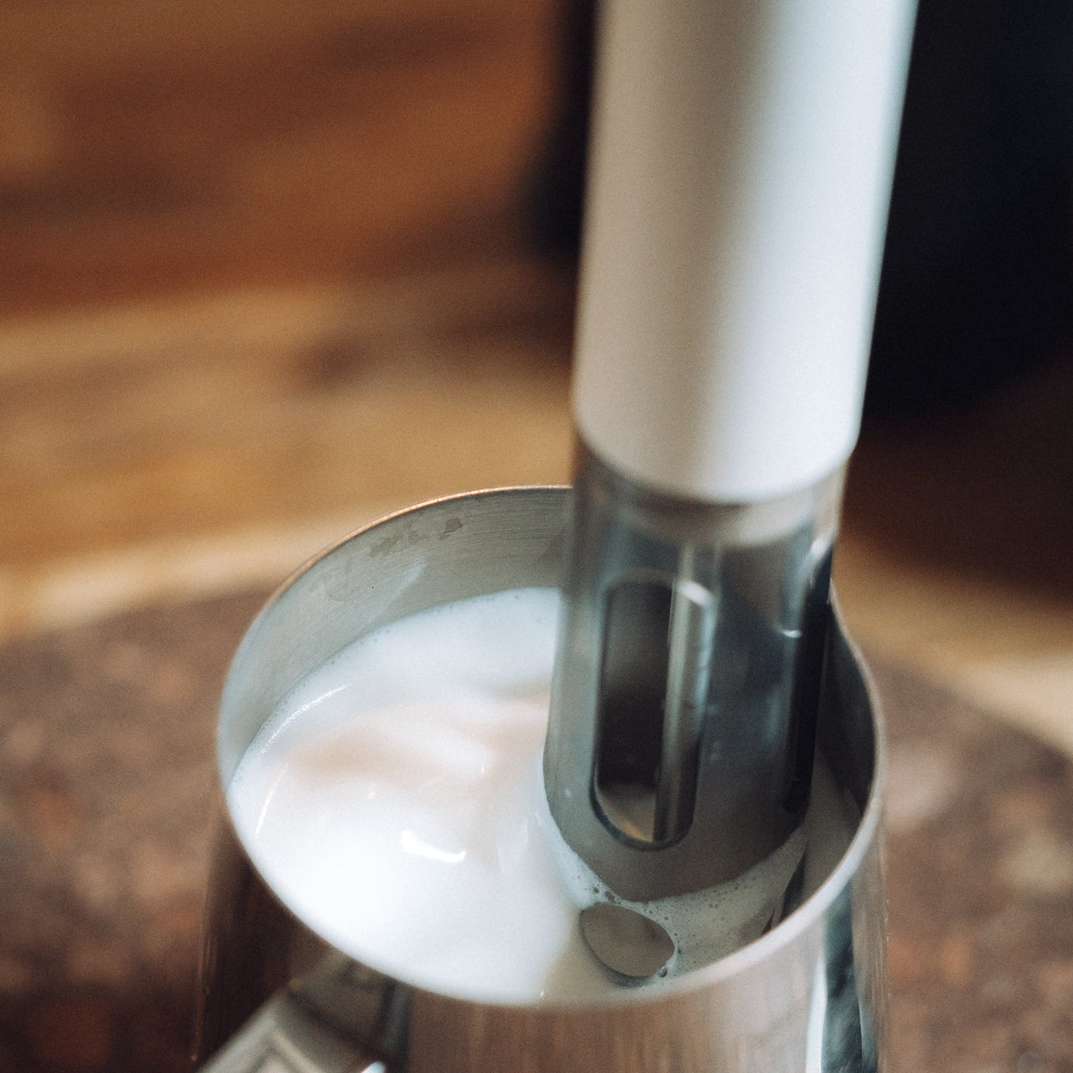 A close-up of a white and stainless steel handheld milk frother being used to foam milk inside a stainless steel pitcher on a wooden surface.