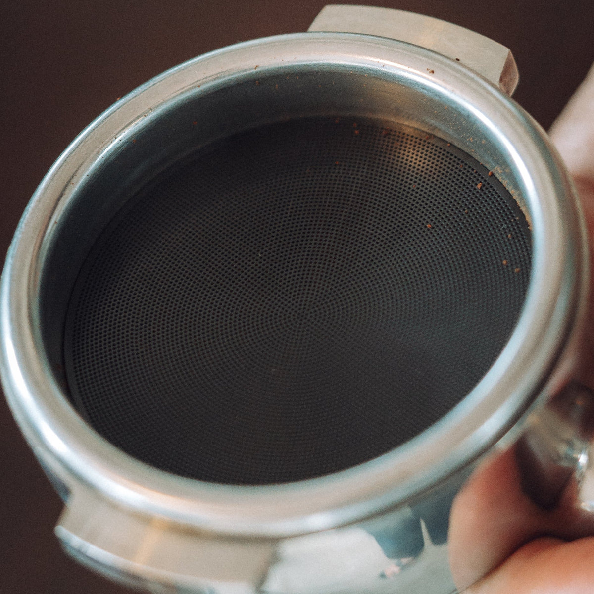 A close-up of a stainless steel portafilter filter basket held by a hand against a dark brown background.