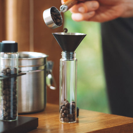 A close-up lifestyle shot of coffee preparation, showing a hand pouring dark coffee beans from a metal scoop into a clear glass carafe with a black funnel, set on a wooden table.