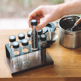 A hand holds a clear glass vial with a black lid containing coffee beans from a black plastic organizer tray on a wooden table, with a metal pot in the background.