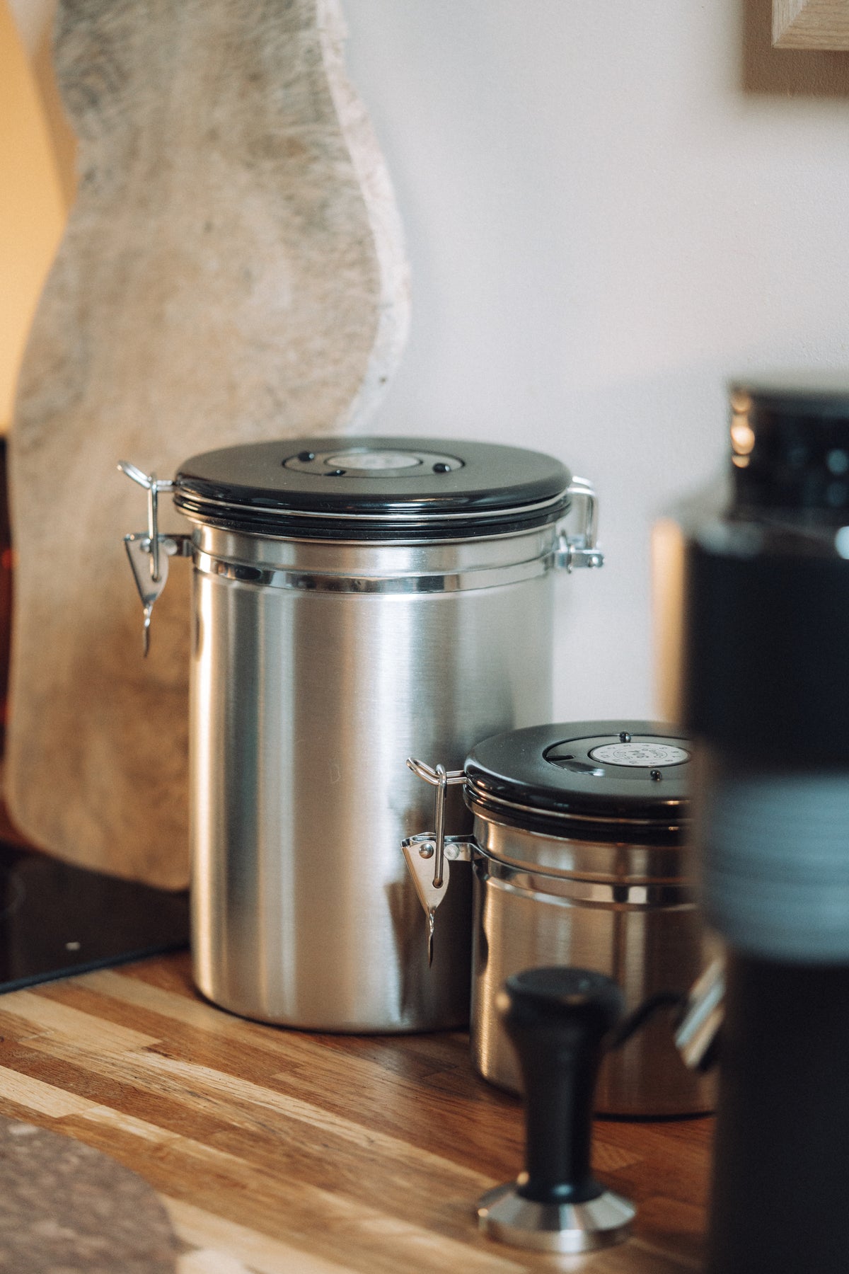 Two stainless steel canisters with black lids, one tall and one short, sitting on a wooden countertop in a kitchen setting.