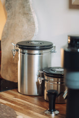 Two stainless steel canisters with black lids, one tall and one short, sitting on a wooden countertop in a kitchen setting.