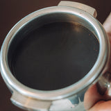 A close-up of a stainless steel portafilter filter basket held by a hand against a dark brown background.