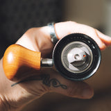 A close-up of a hand holding a vhand coffee grinder with a wooden handle and a silver dials mechanism against a blurred background.