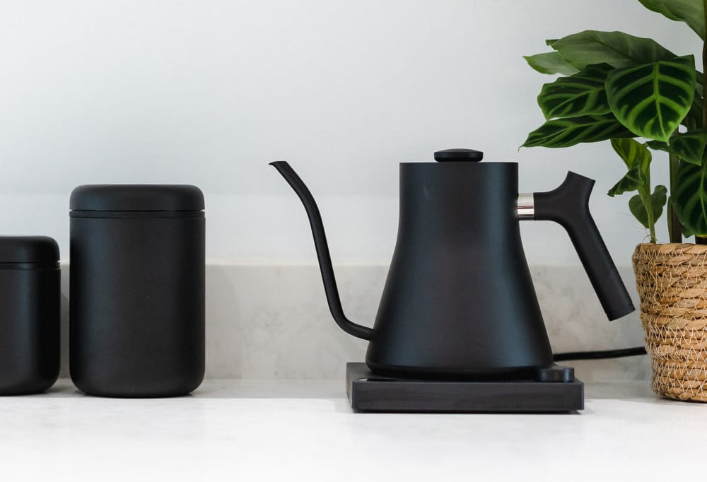 A stylish black Fellow Stagg Kettle sitting on a white counter top, with black coffee storage containers and a plant.