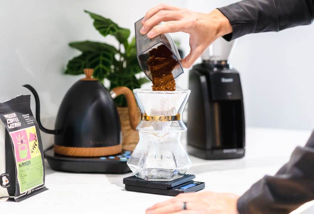 A hand pouring ground coffee into a coffee dripper. Modern, stylish kettle, coffee grinder, packet of coffee and plant are sitting on a white counter top. 