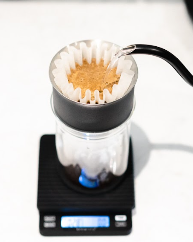 hot water being poured into a bed of ground coffee with a filter paper. the pourer set up is sitting on scales to weigh the water